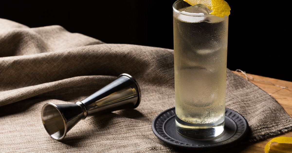 A refreshing drink in a frosty highball glass on on a black coaster. The drink has a slice of lemon peel in it.