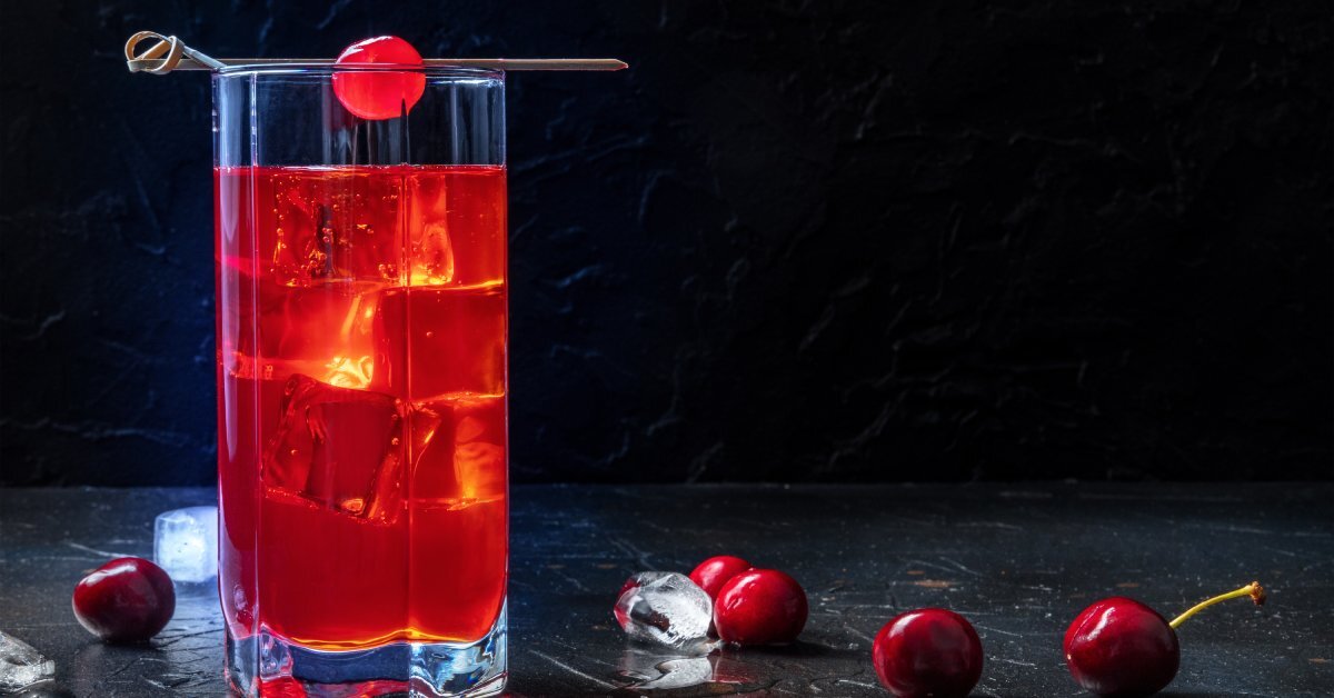 A red-colored iced cocktail in a highball glass on on a table. It’s garnished with a cherry; other cherries are scattered nearby.